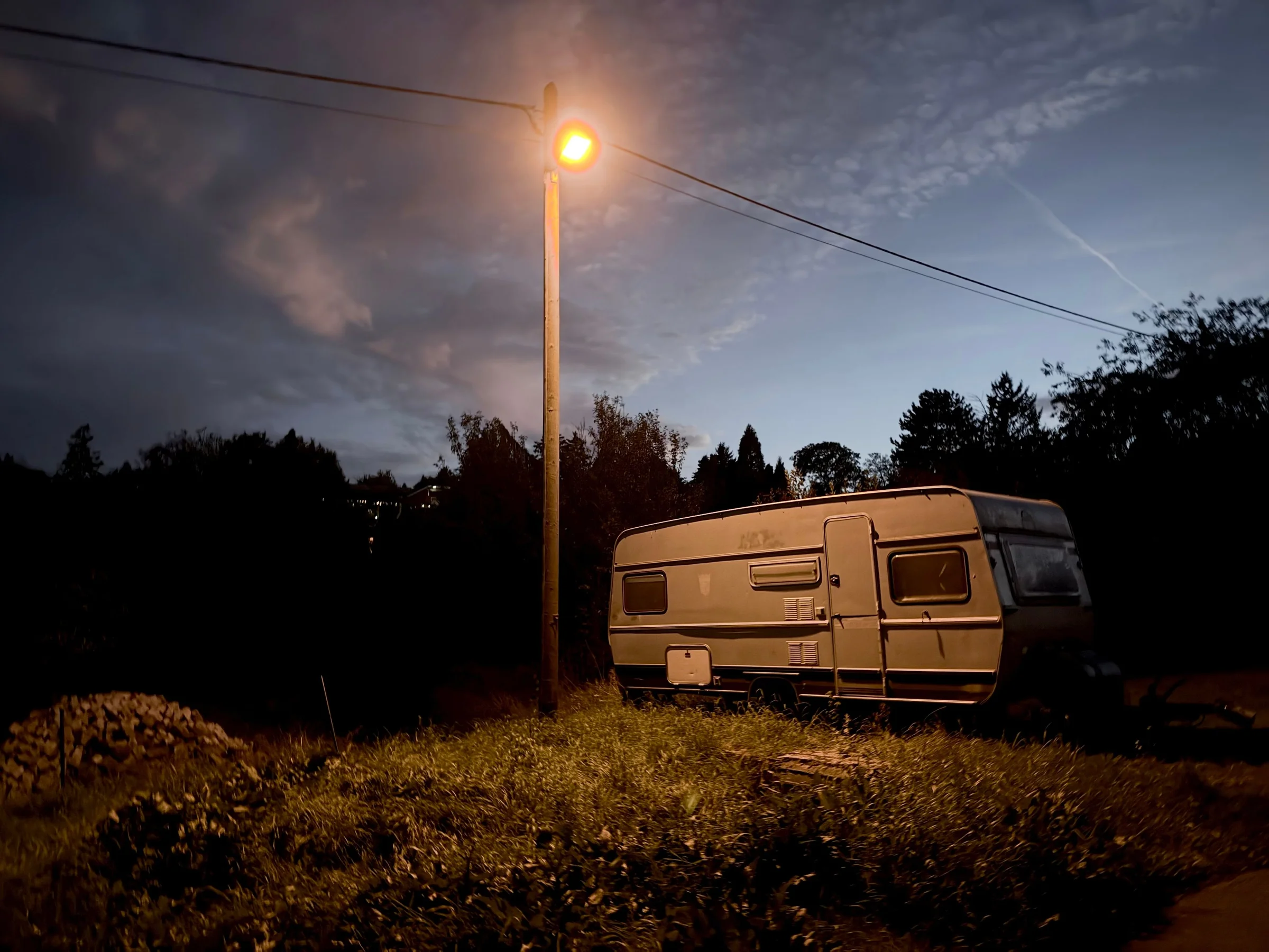 Ein alter, heller Wohnwagen steht in hohem Gras am Rand einer Wiese. Links davon ein Holzmast mit einer Straßenlaterne, die orangefarbenes Licht auf Wagen und Gras wirft. Der Himmel ist dämmerblau mit feinen Wolken, im Hintergrund dunkle Bäume und ein paar beleuchtete Fenster am Hang.